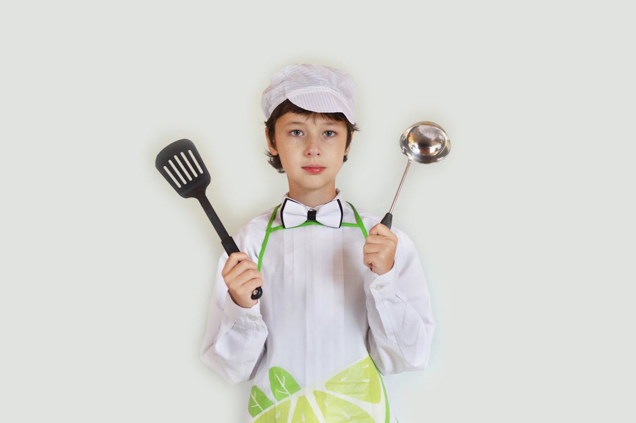 Positive preteen boy in apron and chef hat demonstrating utensils and looking at camera against white wall in studio