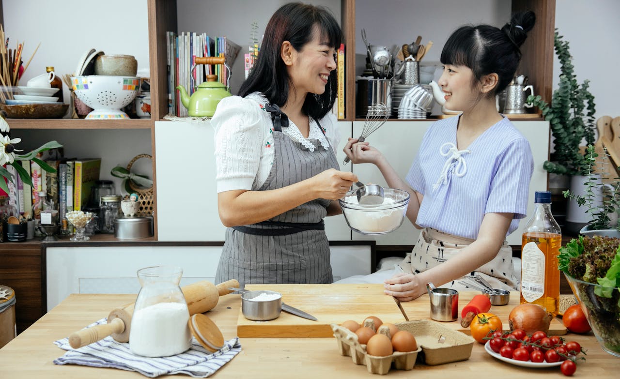 our-story Happy Asian mother wearing apron and daughter standing near wooden counter with ingredients while discussing recipe of pizza