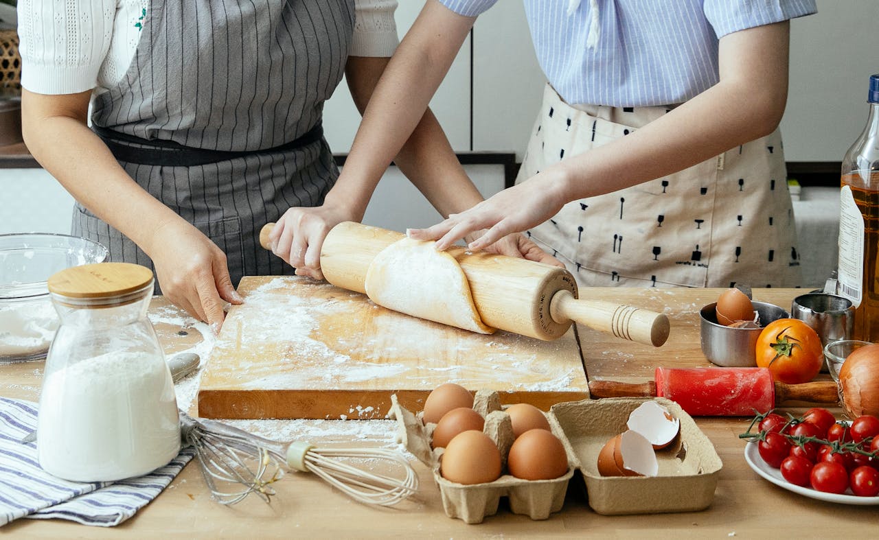 about-us Two women rolling dough together in a home kitchen with fresh ingredients.